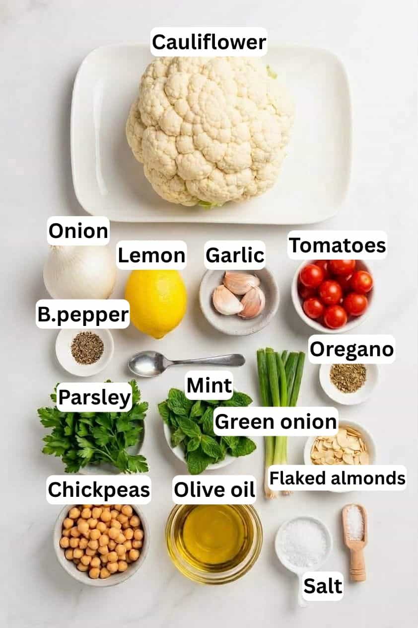 Top view of labeled ingredients on a white surface: cauliflower, onion, lemon, garlic, tomatoes, black pepper, oregano, parsley, mint, green onion, flaked almonds, chickpeas, olive oil, and salt.