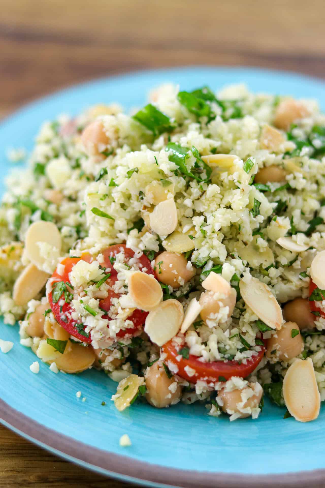 A close-up of a colorful cauliflower salad with cauliflower rice, chickpeas, cherry tomatoes, parsley, and sliced almonds on a blue plate.