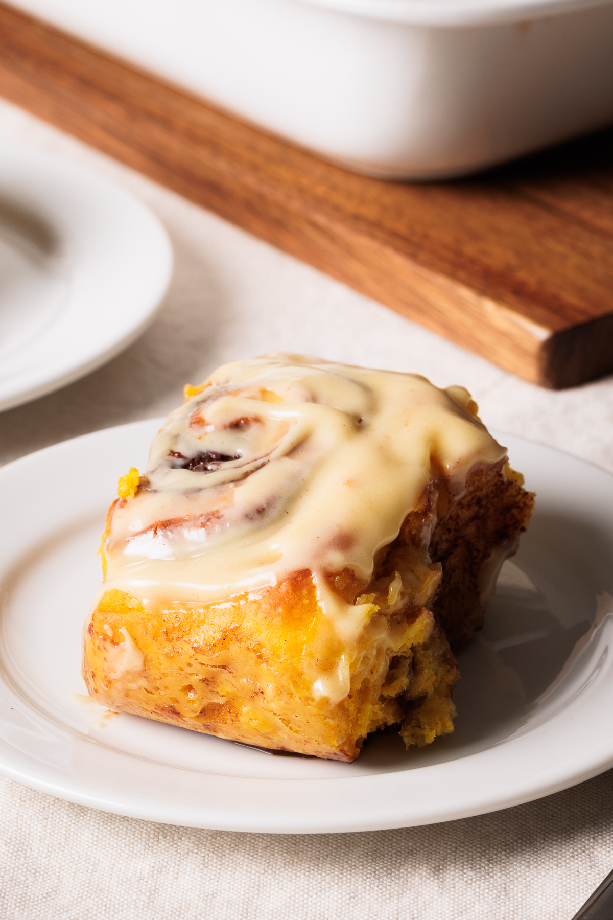 A close-up of a cinnamon roll with pumpkin topped with creamy icing sits on a white plate, with a baking dish and another plate blurred in the background.