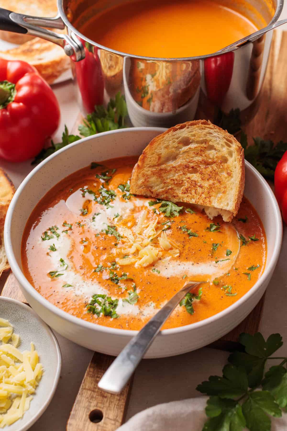 A bowl of Roasted red pepper and gouda soup topped with grated cheese and herbs, served with a slice of toasted bread. A spoon rests in the bowl, and fresh tomatoes and a pot of soup are in the background.