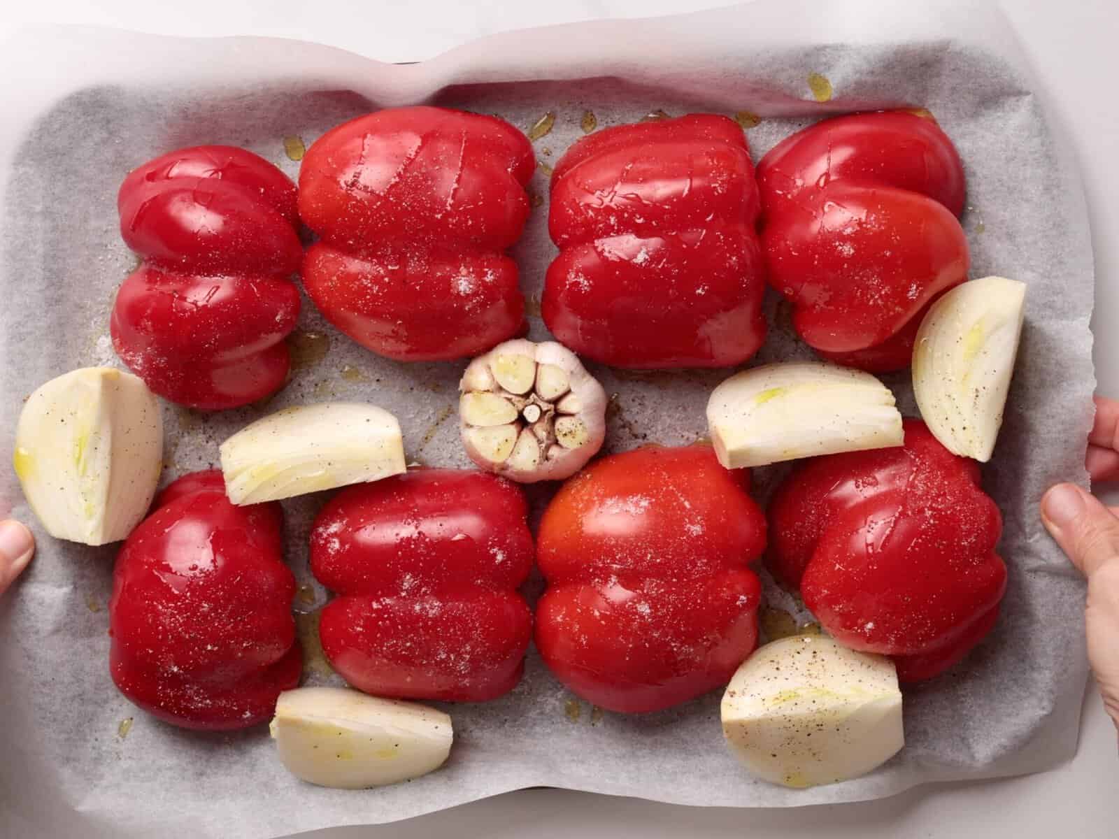Red bell peppers, quartered onions, and a halved garlic head are arranged on a parchment-lined baking tray, sprinkled with salt, pepper, and drizzled with oil, ready for roasting.