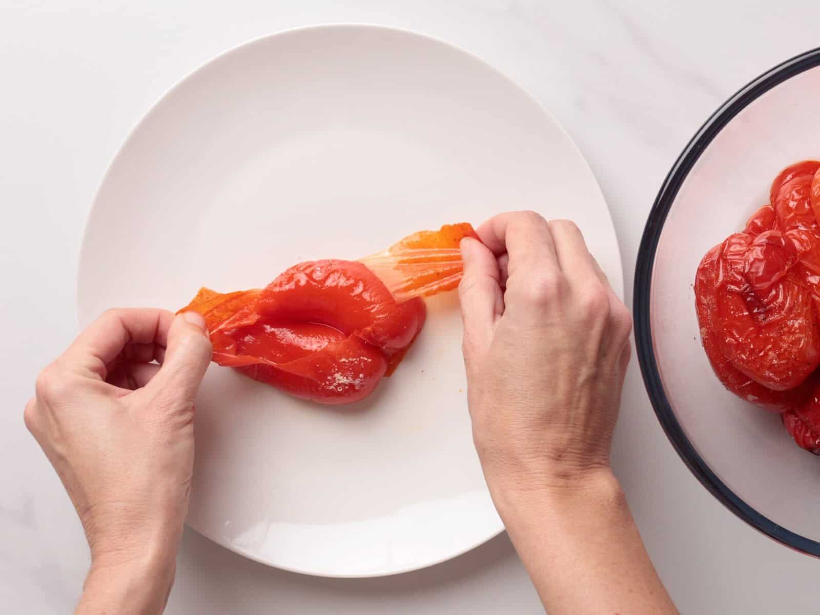 Two hands peeling the skin off a red roasted pepper on a white plate, with more peeled peppers visible in a glass bowl to the side.