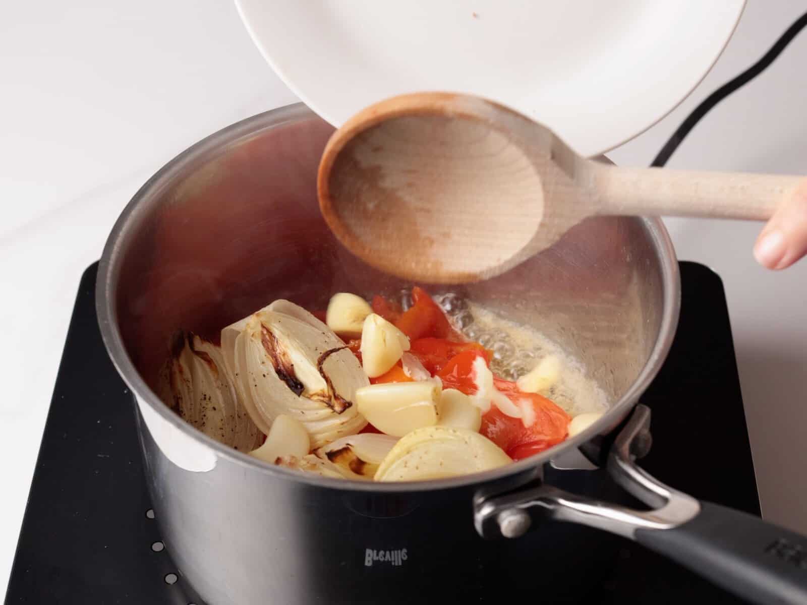 A hand holds a wooden spoon and tilts a plate, adding roasted vegetables—onion, garlic, and peppers—into a pot on a stove.