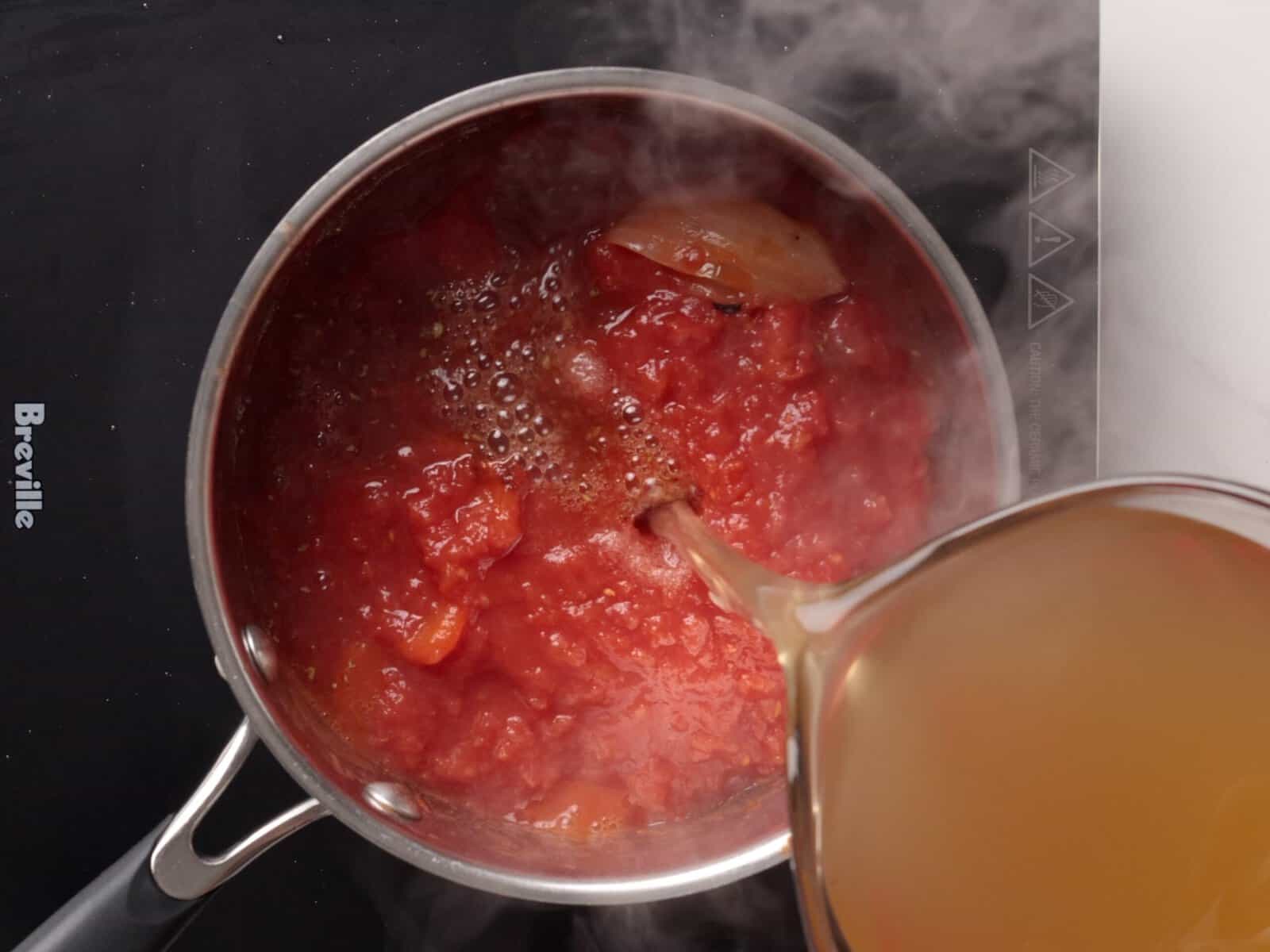 A hand pours broth from a measuring cup into a pot of simmering tomato sauce on a black stovetop, with steam rising from the pot.