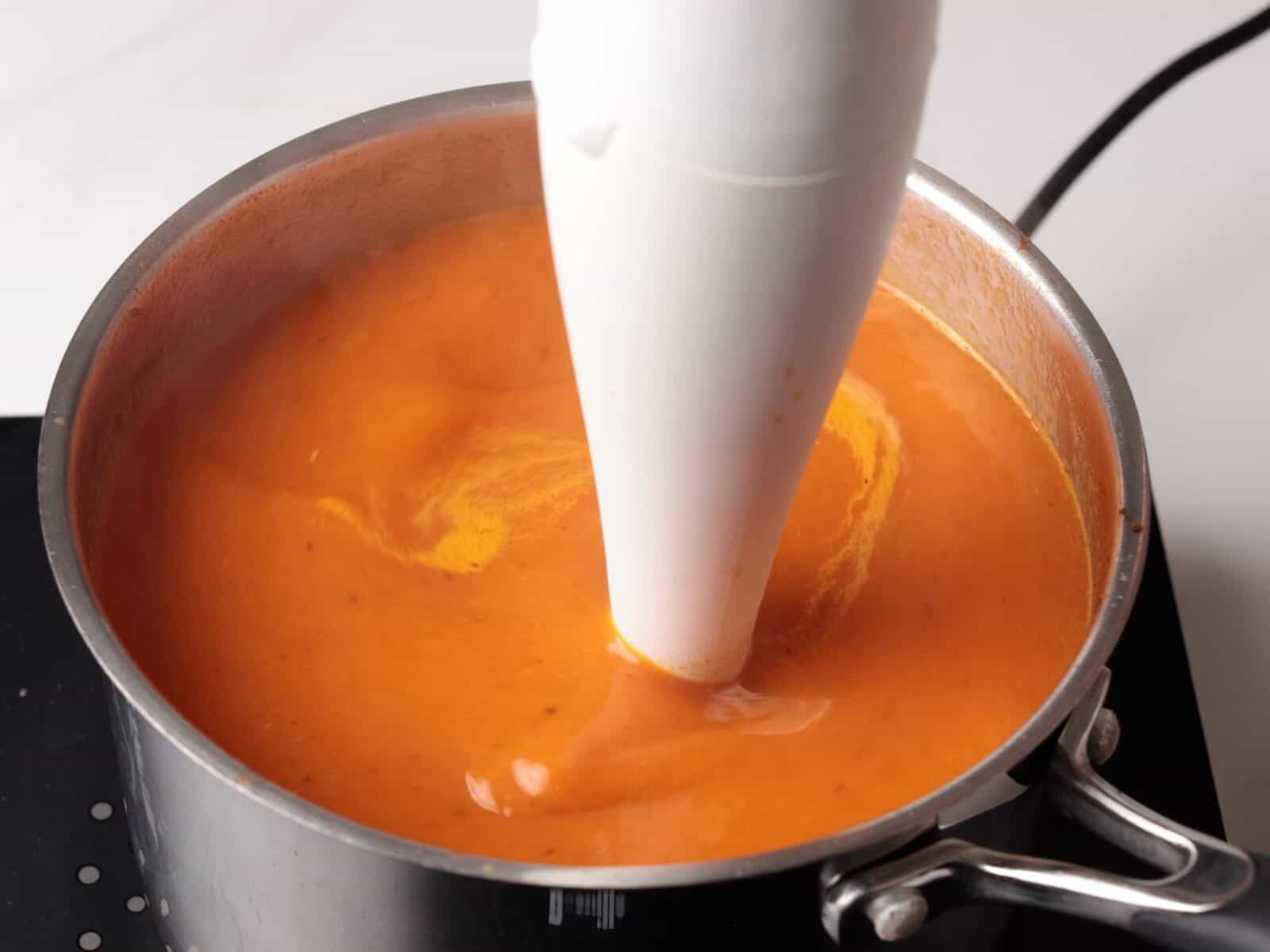 An immersion blender is blending orange soup inside a metal pot on a stovetop. The soup appears creamy and smooth.