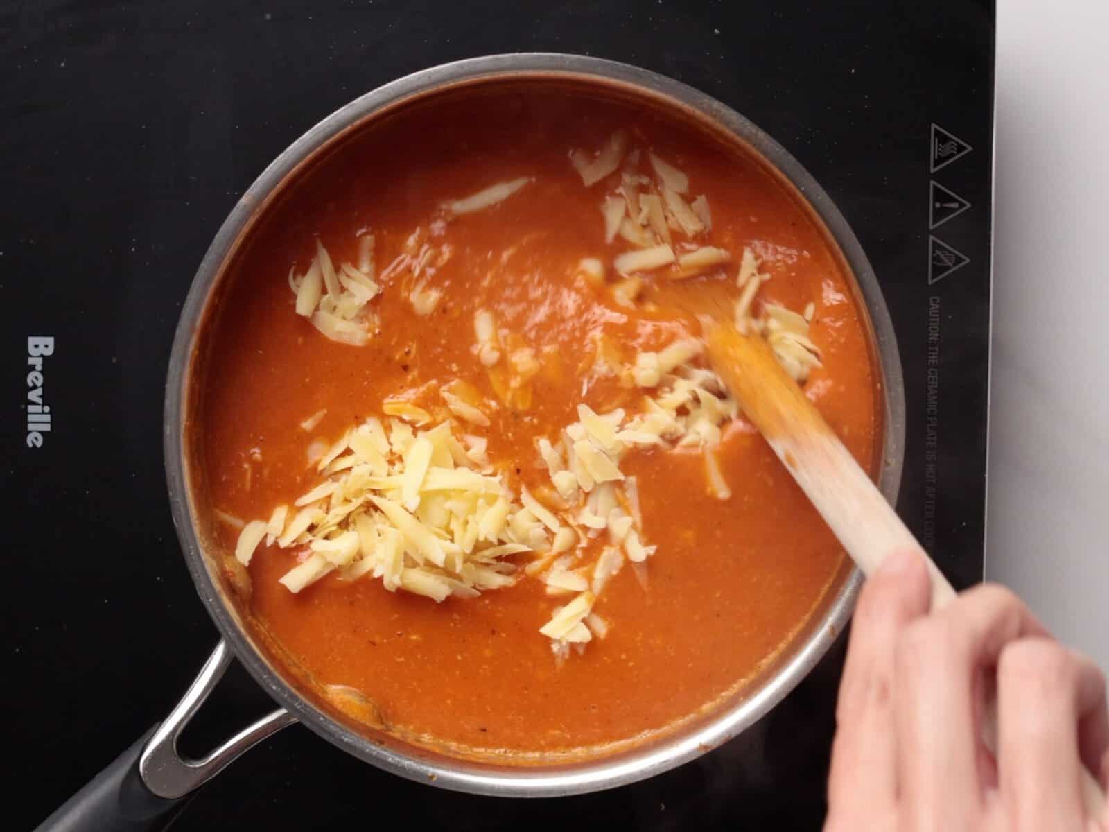 A hand stirs shredded Gouda cheese into a pot of orange tomato soup on a black stovetop.