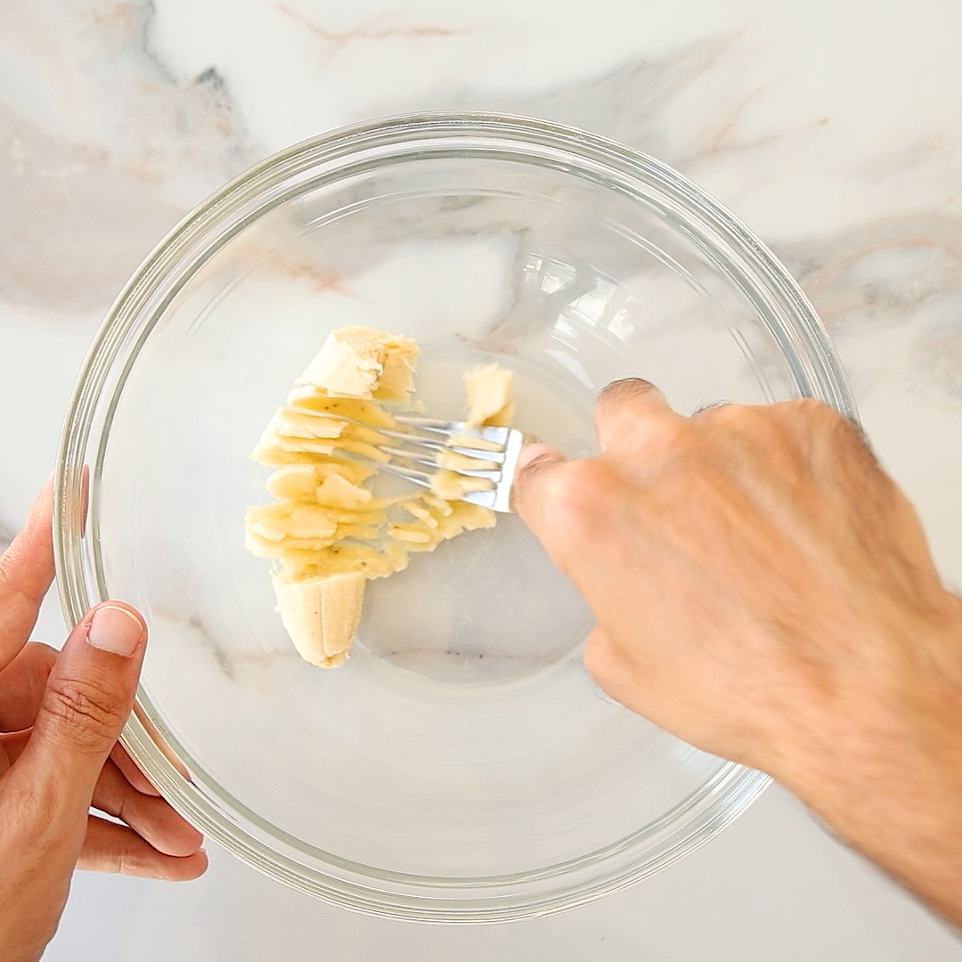 A person uses a fork to mash a banana in a glass bowl.