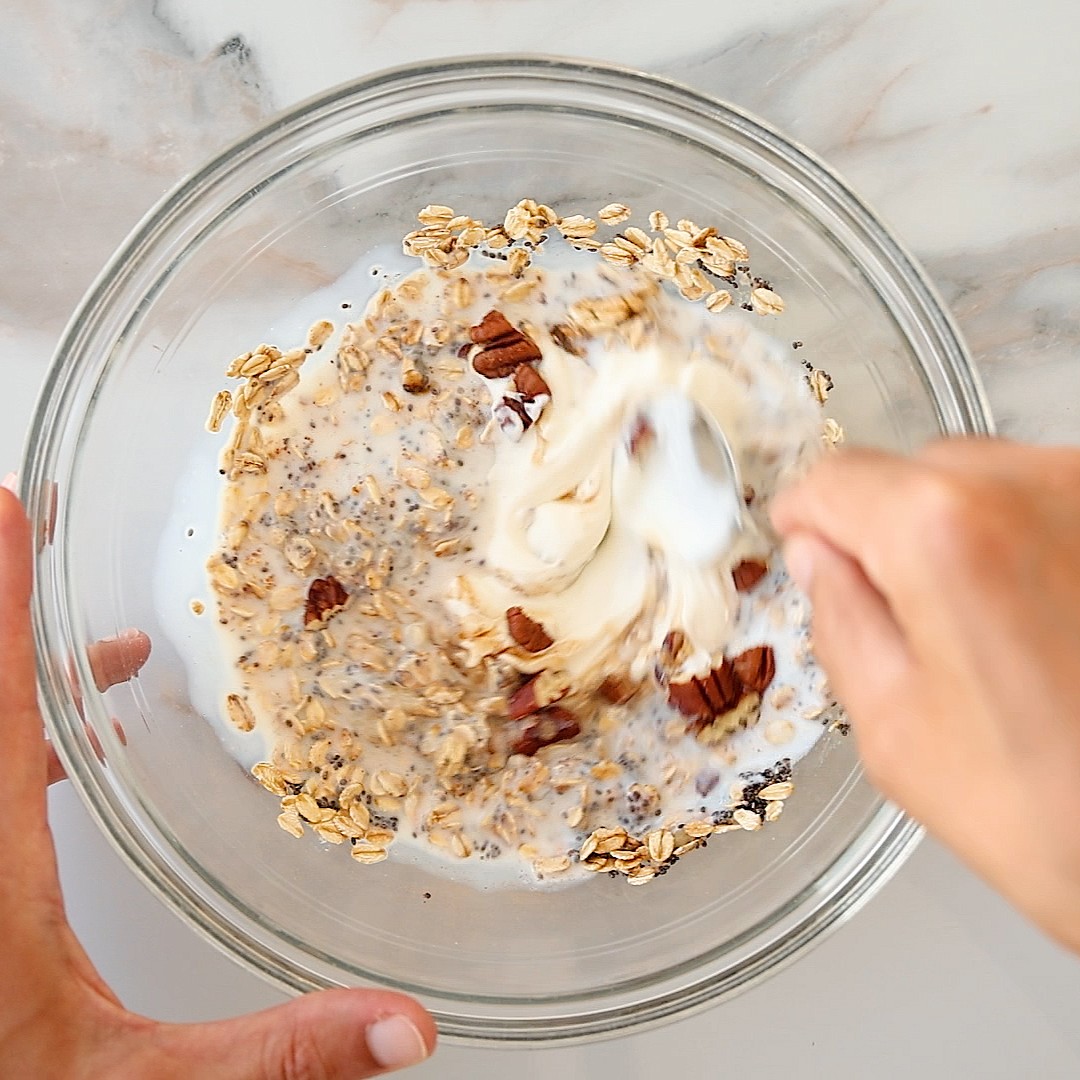 A hand stirs oats, chia seeds, milk, pecans, and a dollop of yogurt in a glass bowl.