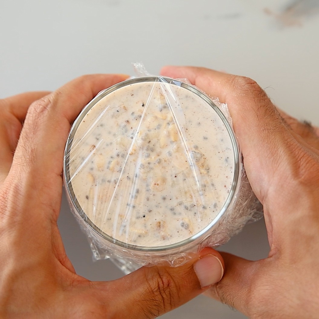 Two hands holding a glass jar filled with overnight oats covered in plastic wrap.