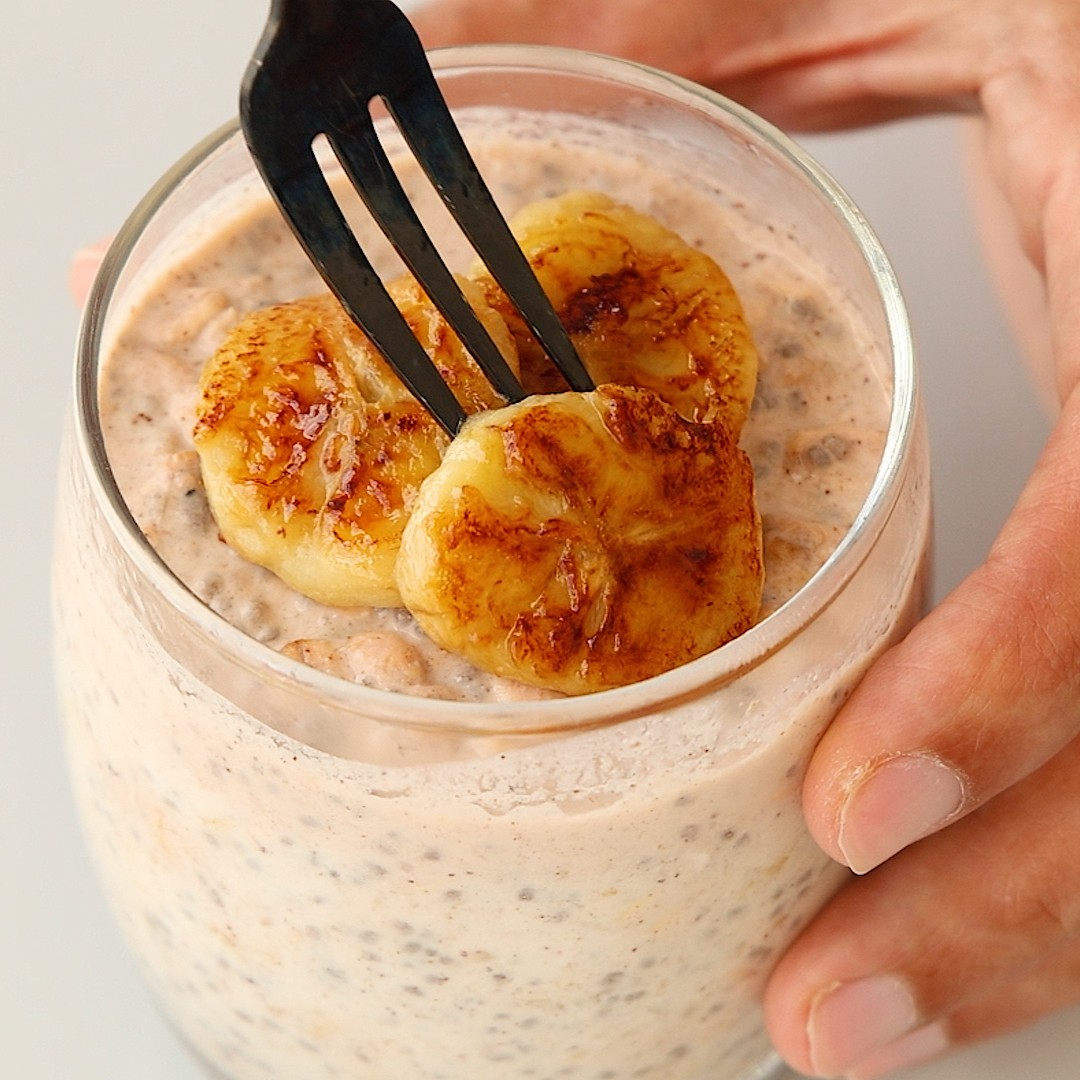 A hand holds a glass of banana bread overnight oats topped with caramelized banana slices being lifted by a fork.