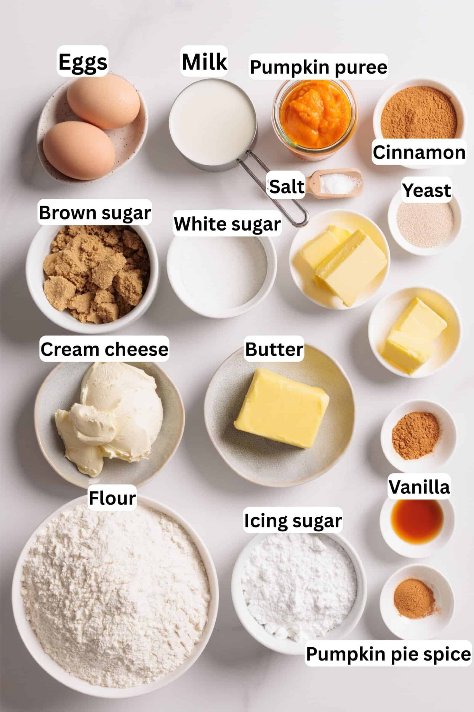 Various baking ingredients for pumpkin cinnamon rolls labeled in bowls on a white surface, including eggs, milk, pumpkin puree, brown and white sugar, cream cheese, flour, butter, icing sugar, salt, cinnamon, yeast, vanilla, and pumpkin pie spice.