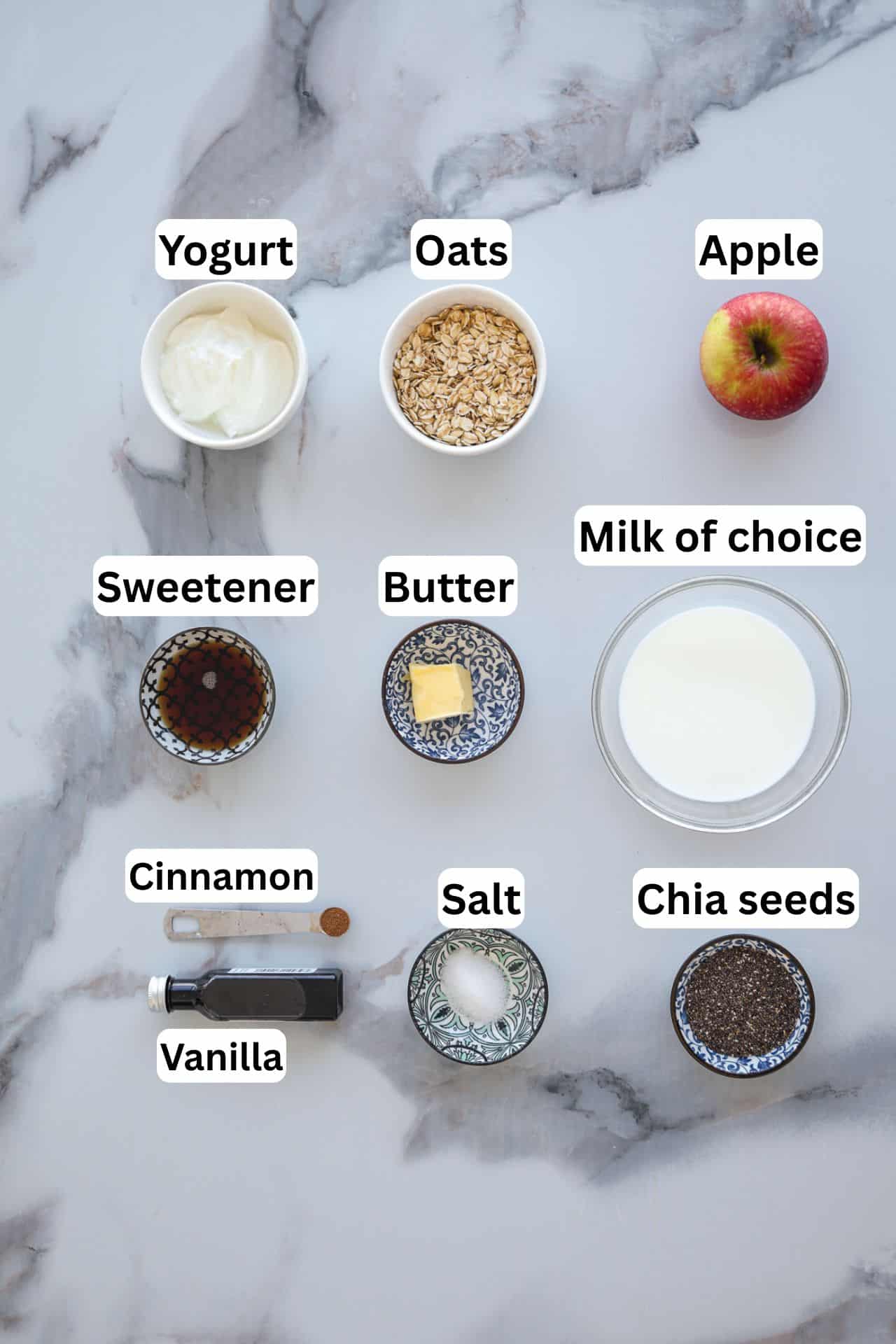 Ingredients for apple pie overnight oats recipe labeled as yogurt, oats, apple, sweetener, butter, milk, cinnamon, vanilla, salt, and chia seeds arranged on a marble surface.