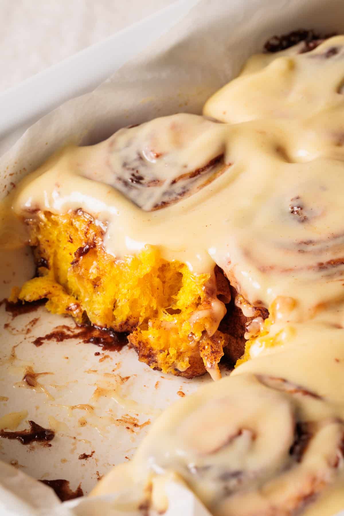 Close-up of PUMPKIN SPICE cinnamon rolls covered in creamy icing, with one roll partially eaten, showing the soft, yellow-orange interior and swirls of cinnamon. The rolls sit in a white, parchment-lined baking dish.
