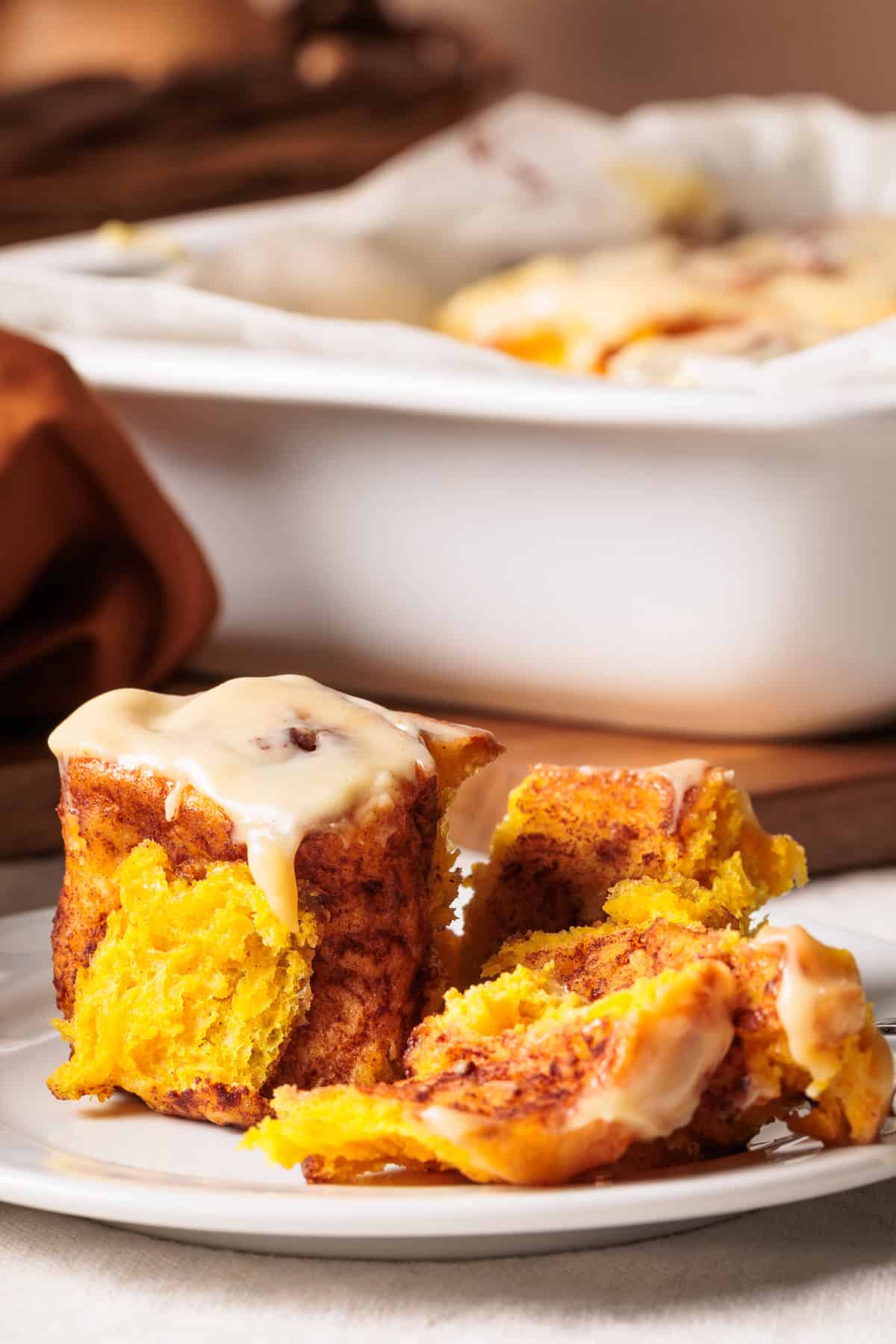 A plate holds one whole and one partially eaten pumpkin cinnamon roll with cream cheese frosting. The soft, bright orange dough is topped with a sweet glaze, sitting before a baking dish filled with more rolls on a wooden surface.