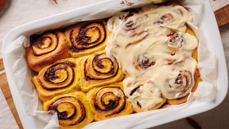 A baking dish with cinnamon rolls, half covered in creamy icing and half without, showing the swirled tops. The rolls sit on parchment paper in a white dish.