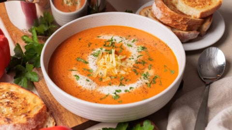 Smoky Roasted Red Pepper Gouda Soup A bowl of creamy roasted red pepper and gouda soup garnished with shredded gouda cheese and herbs, surrounded by toasted bread slices on a plate, fresh parsley, a spoon, and a beige napkin.