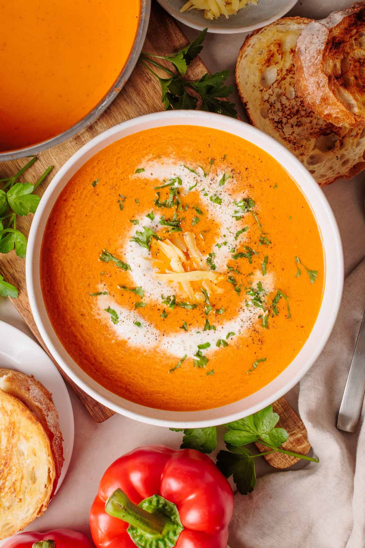 A bowl of roasted red pepper gouda soup garnished with cream, shredded cheese, and chopped herbs, surrounded by bread, parsley, a red bell pepper, and a grilled cheese sandwich.