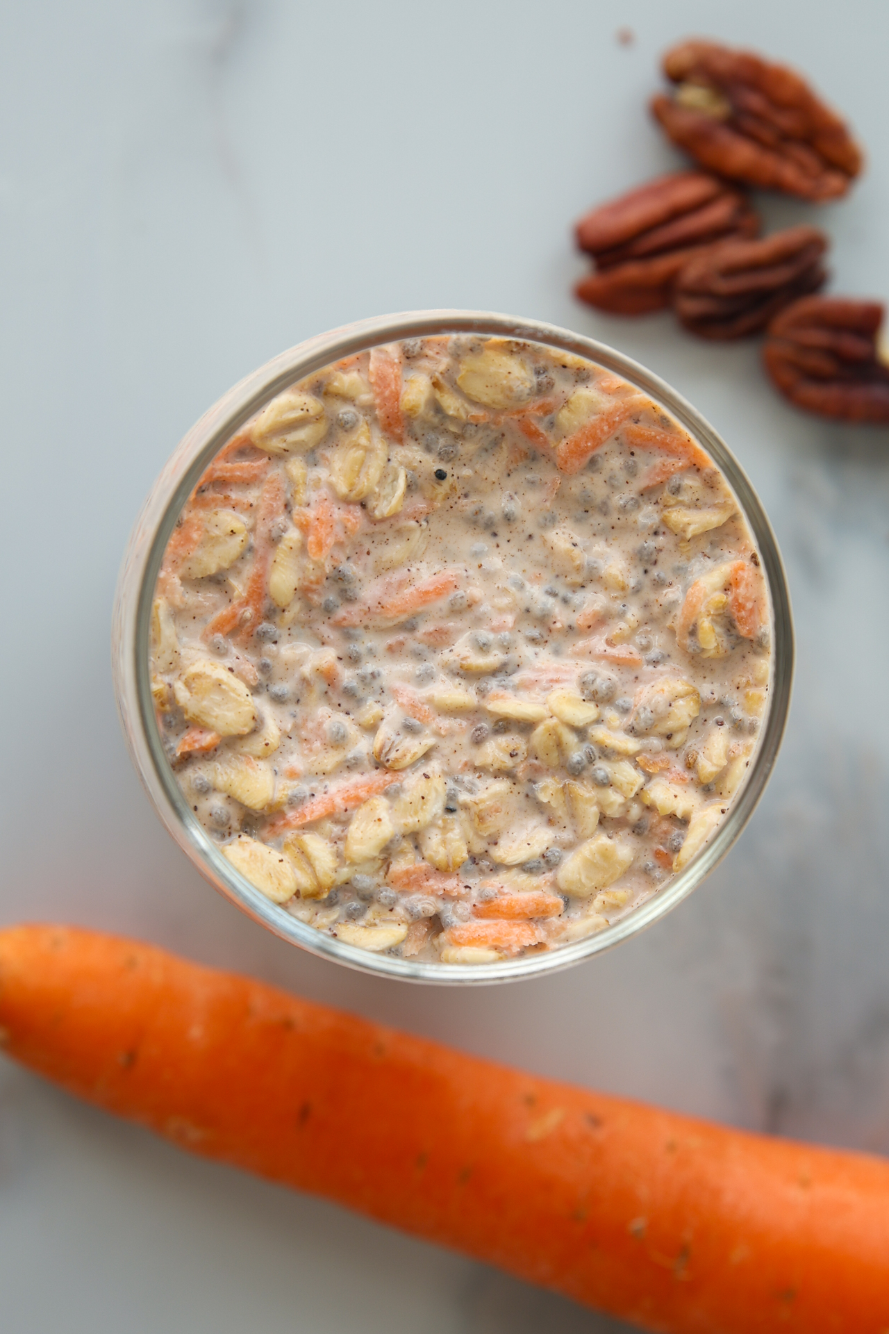 A glass jar of carrot cake overnight oats topped with chia seeds, with a whole carrot and a few pecans nearby.
