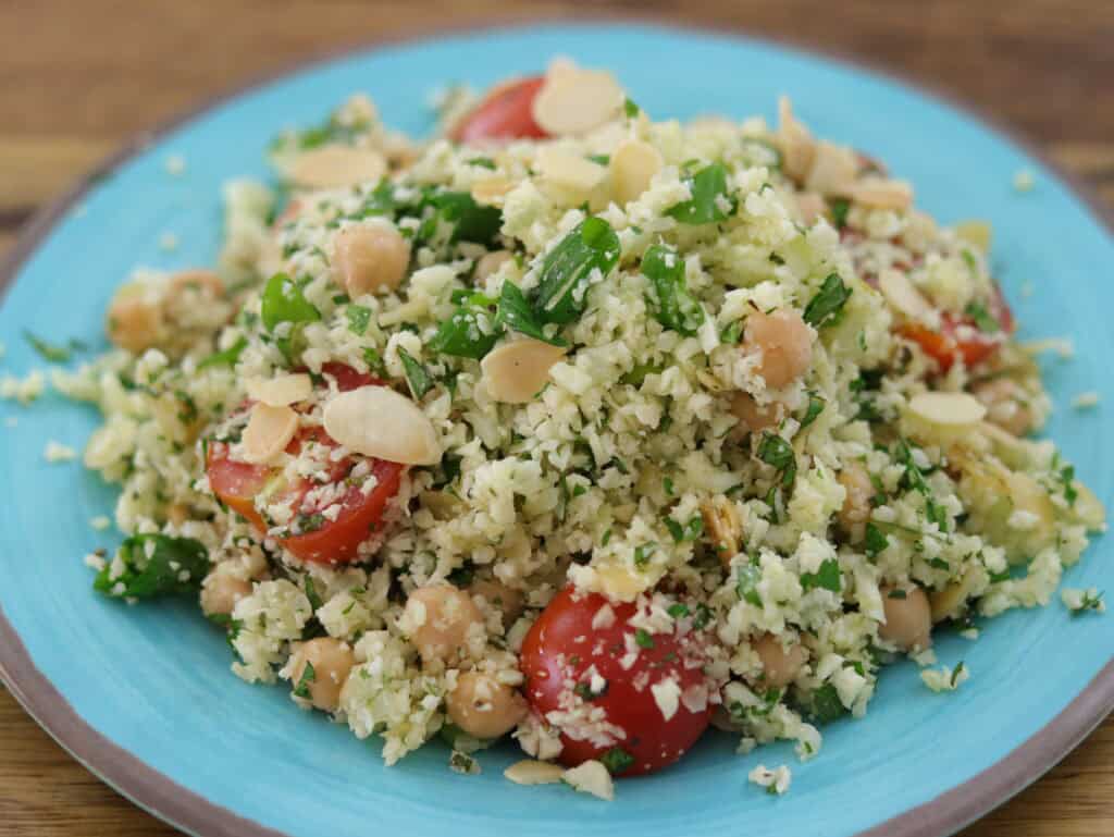 A blue plate with Mediterranean cauliflower rice salad, featuring cherry tomatoes, chickpeas, green herbs, and sliced almonds on a wooden surface.