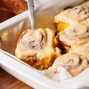 A close-up of pumpkin cinnamon rolls with cream cheese frosting in a white baking dish. One roll is being lifted out with a metal spatula.