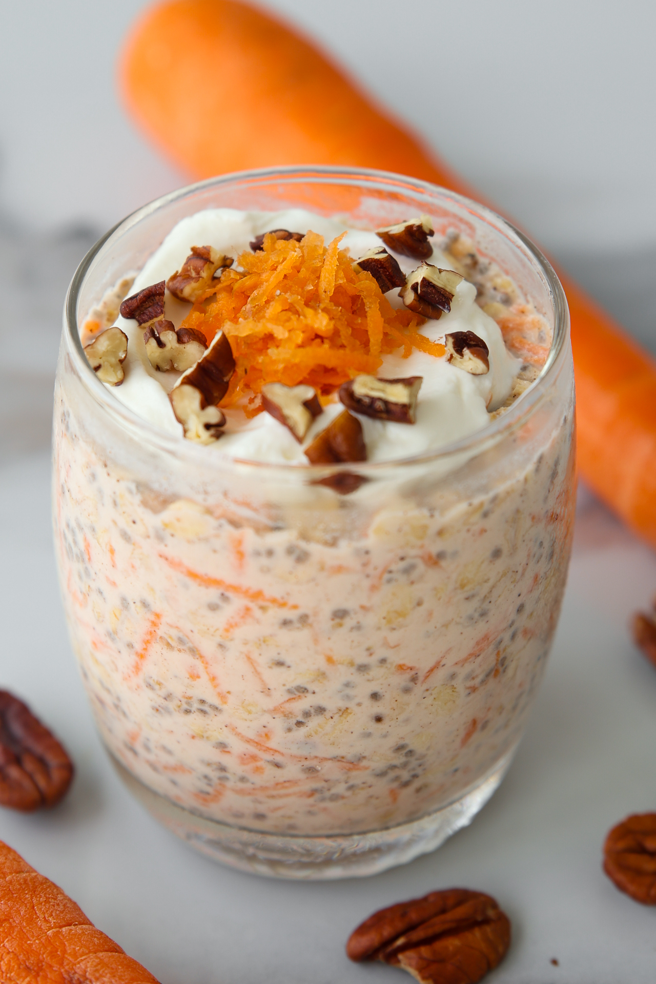 A glass jar of carrot cake overnight oats topped with yogurt, shredded carrots, and pecans, with whole carrots and pecans nearby.