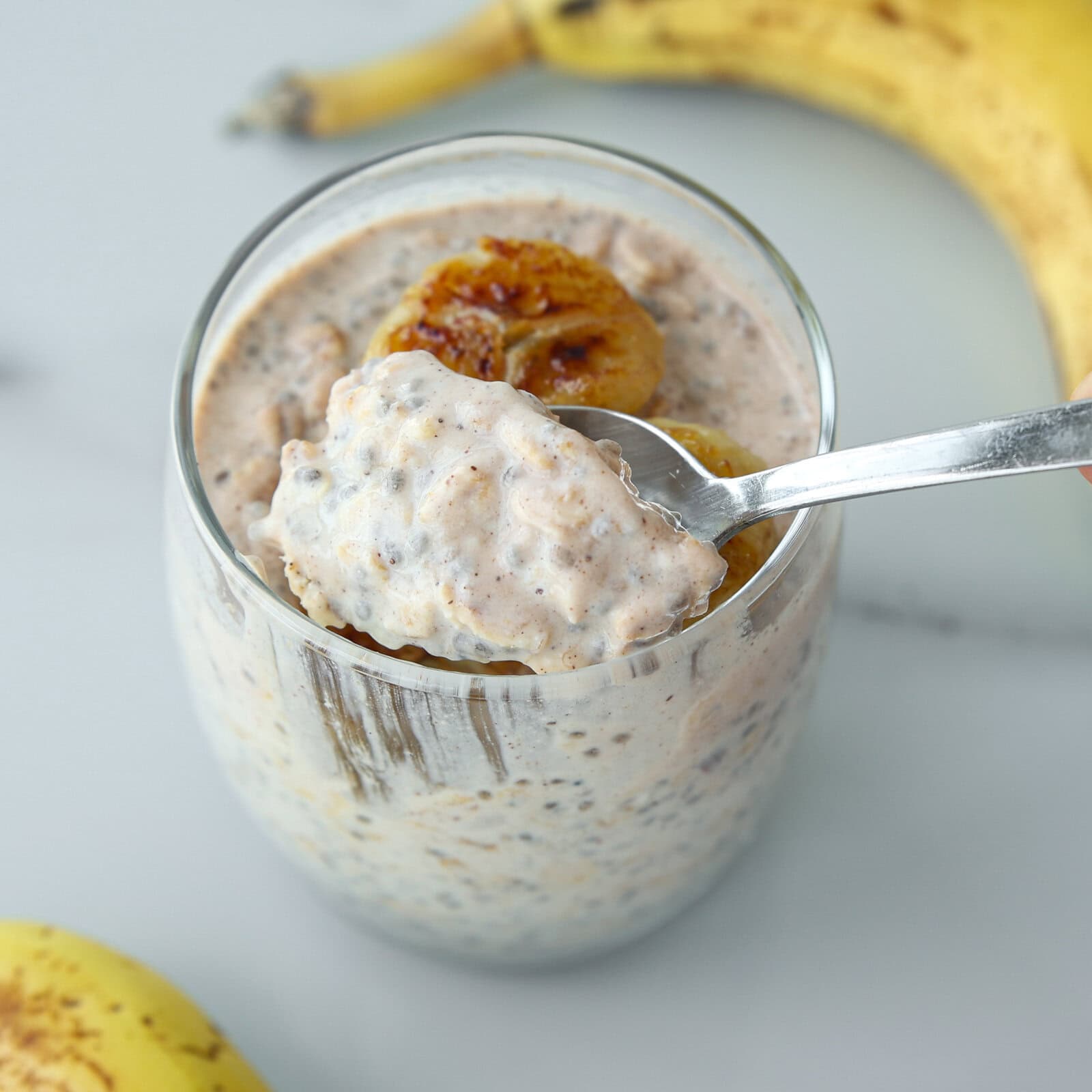 A spoonful of banana bread overnight oats held over a glass, with bananas in the background.