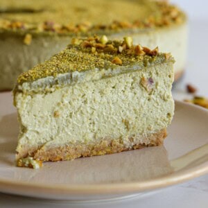 A slice of pistachio cheesecake with a nut crust on a plate, with the rest of the cake in the background.