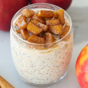 A glass of apple pie overnight oats topped with sautéed cinnamon apples, with fresh apples and a cinnamon stick nearby.