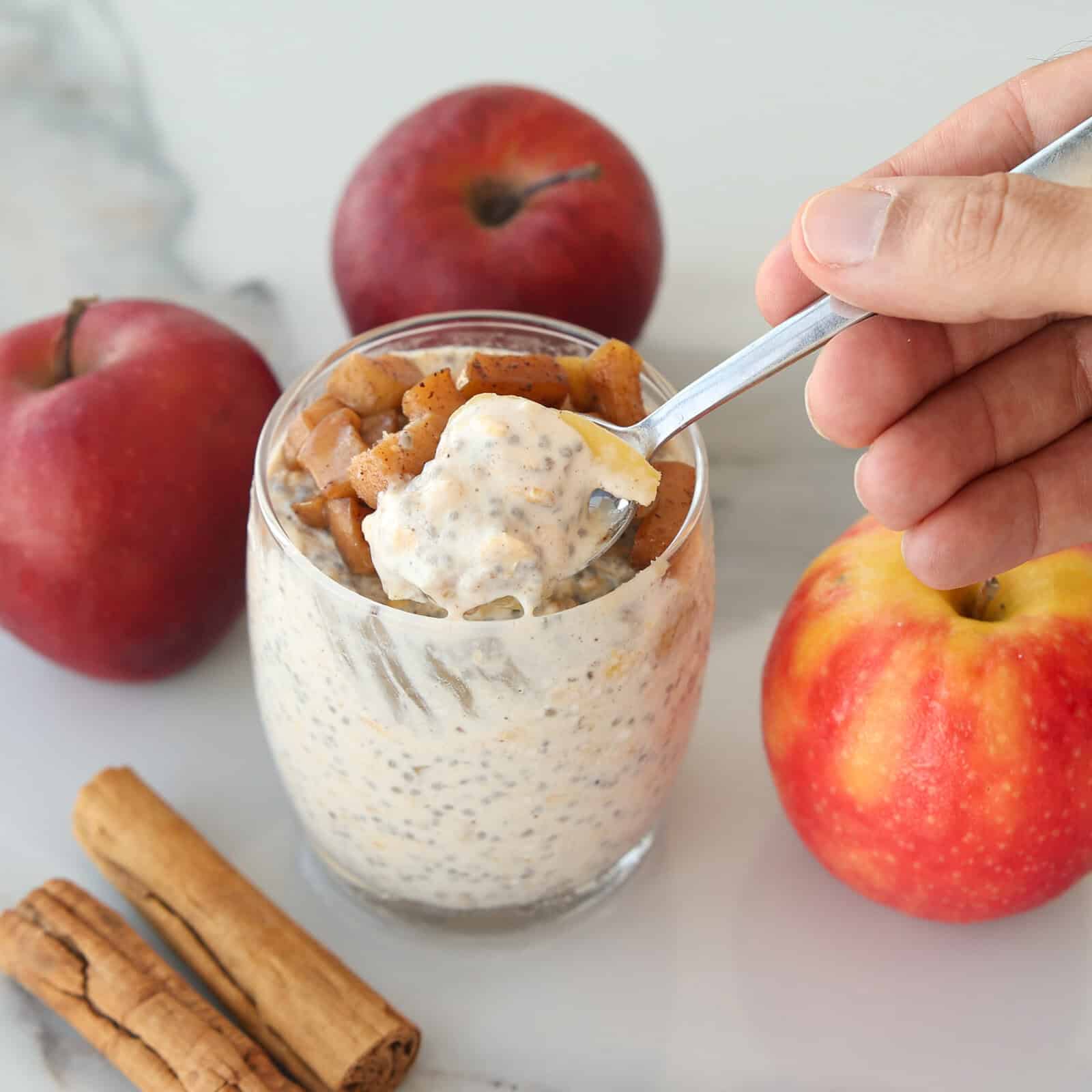A hand holding a spoonful of apple cinnamon overnight oats over a glass jar, surrounded by whole apples and cinnamon sticks.