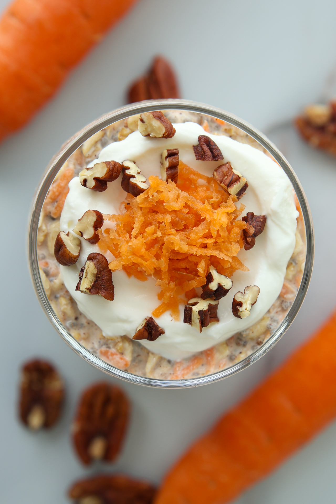 Glass jar of carrot cake overnight oats topped with yogurt, shredded carrots, and pecans, surrounded by whole carrots and pecans.