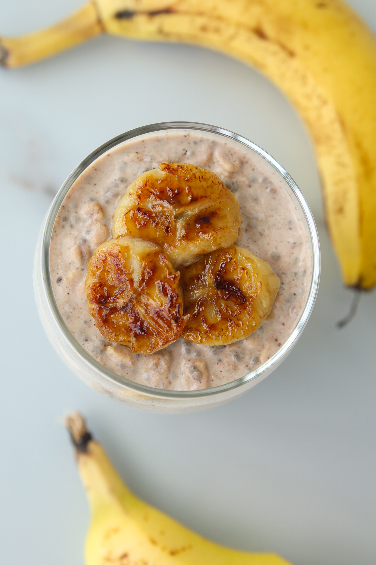 A glass of banana overnight oats topped with three slices of caramelized banana, with two whole bananas nearby.