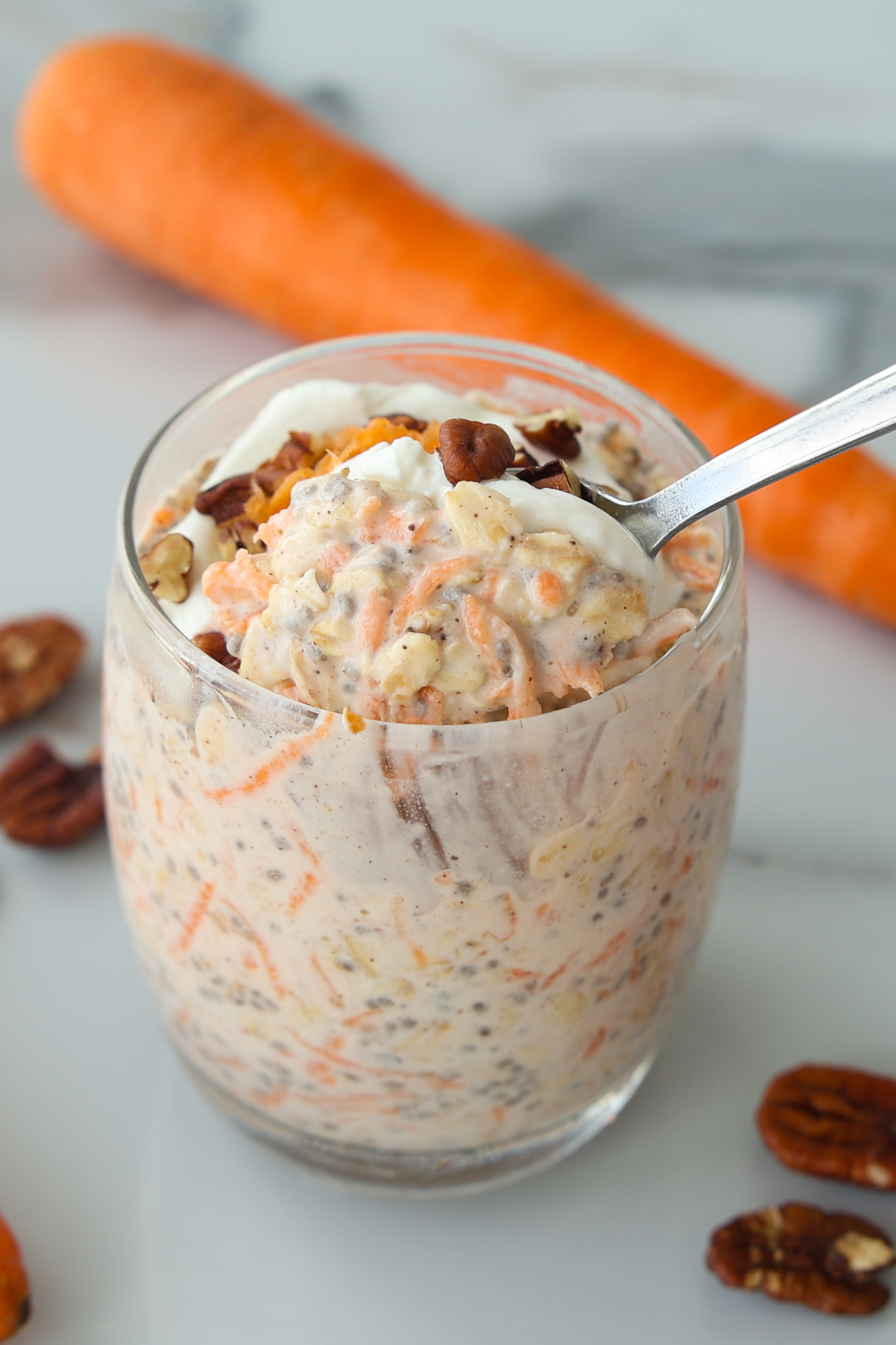 A glass of carrot cake overnight oats topped with pecans and a dollop of yogurt, with a spoon lifting a portion.
