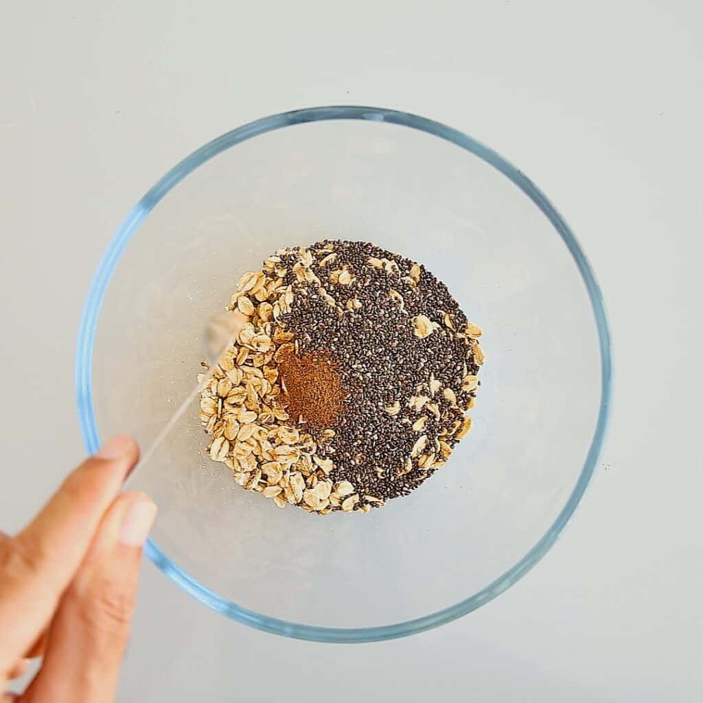 A hand holds a spoon over a clear glass bowl containing rolled oats, chia seeds, and a spoonful of ground cinnamon.