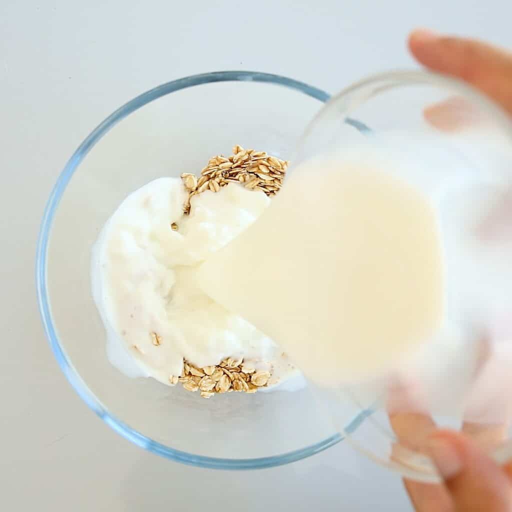 Milk being poured from a small glass into a bowl containing yogurt and oats.