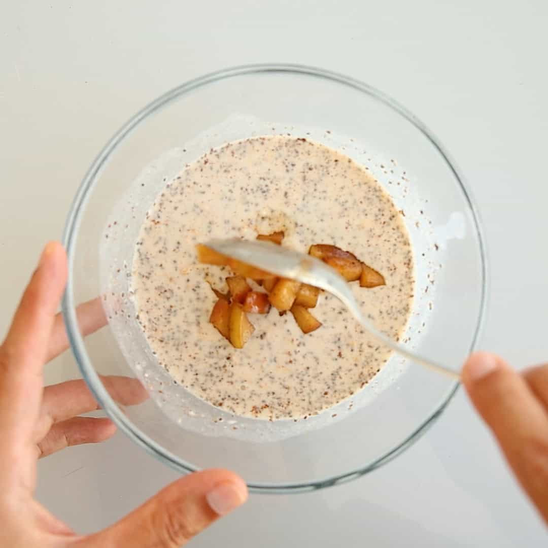 A spoon adding cooked apple slices to a glass bowl of creamy overnight oats mixture with chia seeds.
