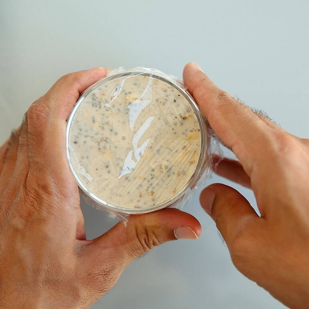 Two hands hold a small, round container filled with a creamy, overnight oats mixture covered in plastic wrap.