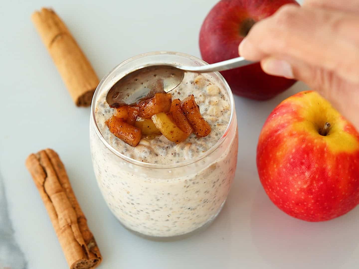 A hand uses a spoon to top a glass of overnight oats with cooked cinnamon apples, surrounded by two whole apples and cinnamon sticks.