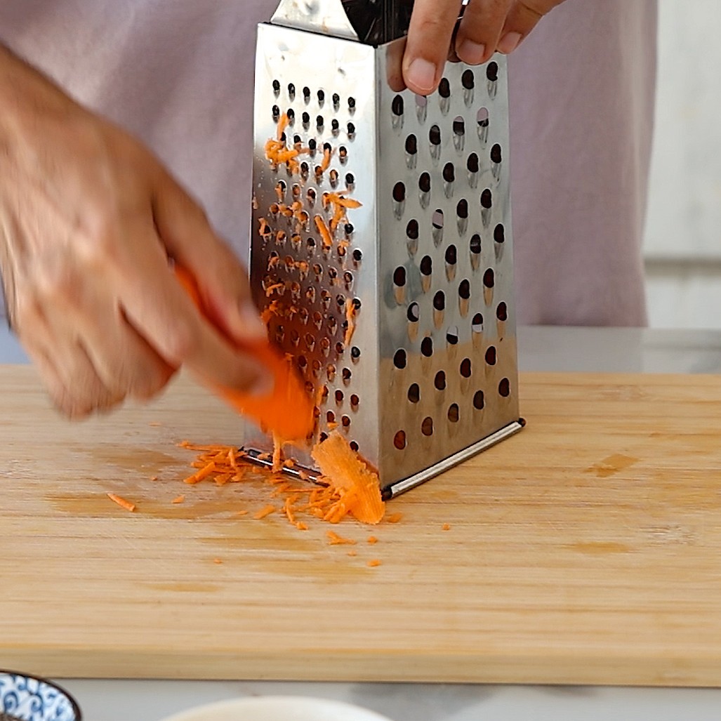 Hands grating a carrot on a metal box grater over a wooden cutting board.