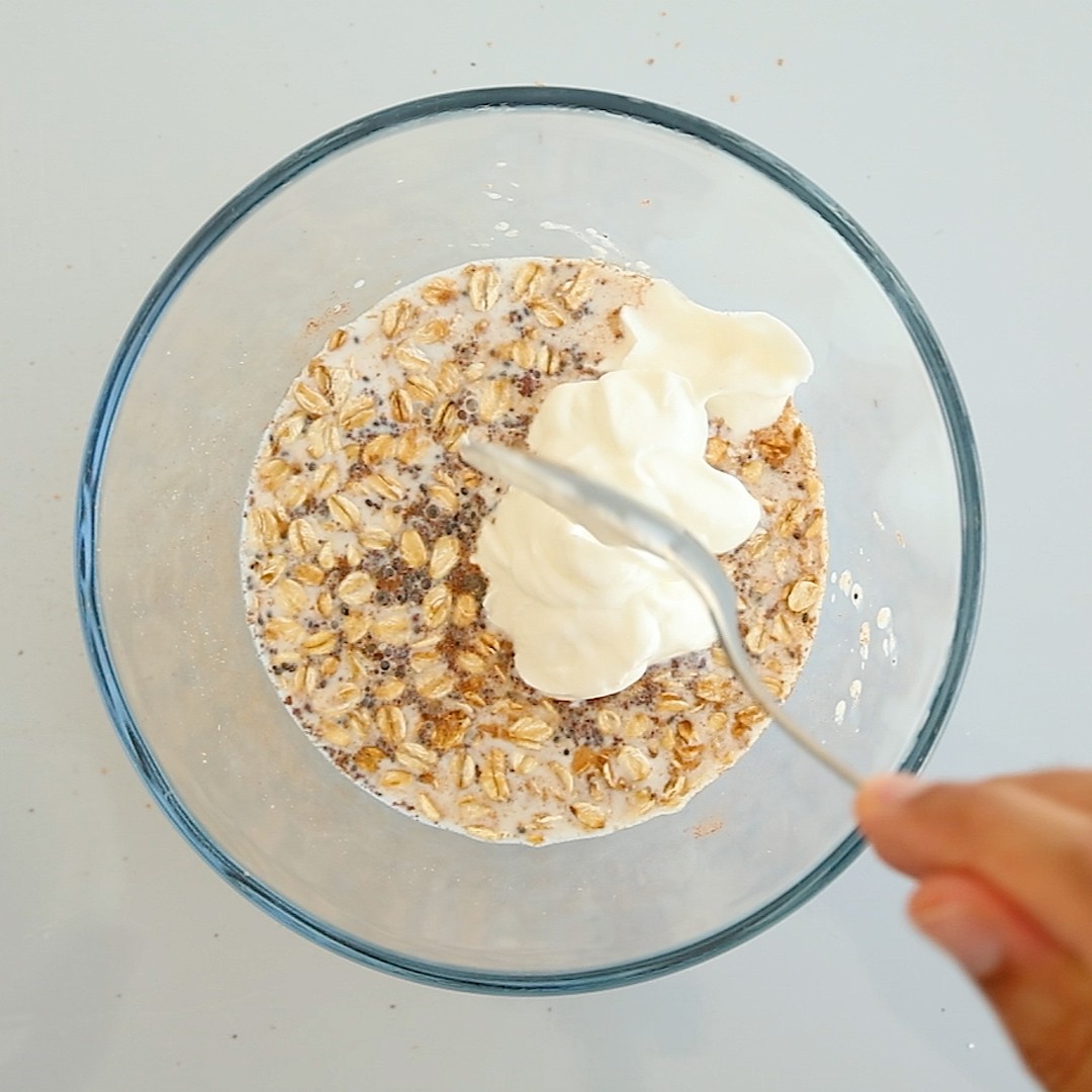 A hand holding a spoon mixes Greek yogurt into a bowl of oats and chia seeds.
