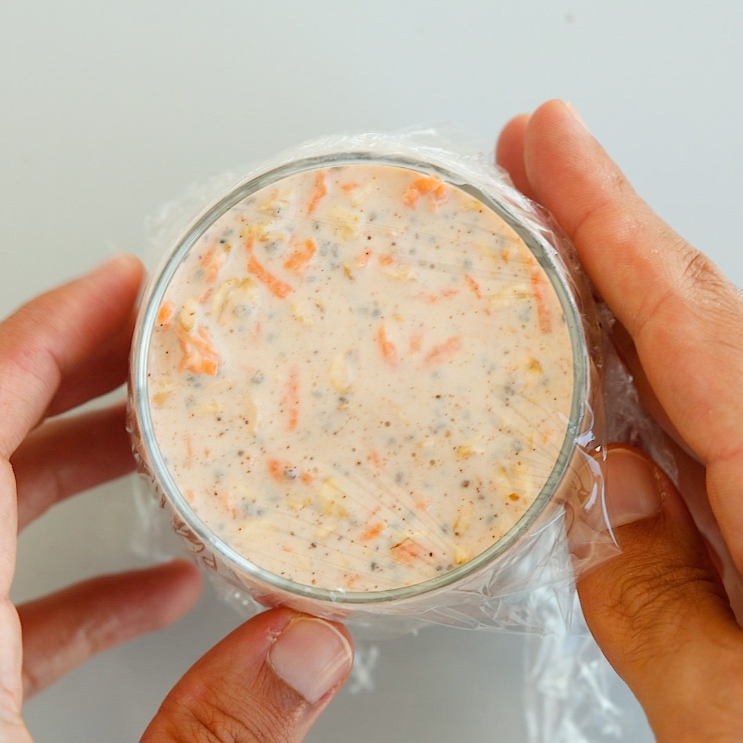 Two hands holding a glass container filled with a creamy carrot and spice overnight oats, covered with plastic wrap.