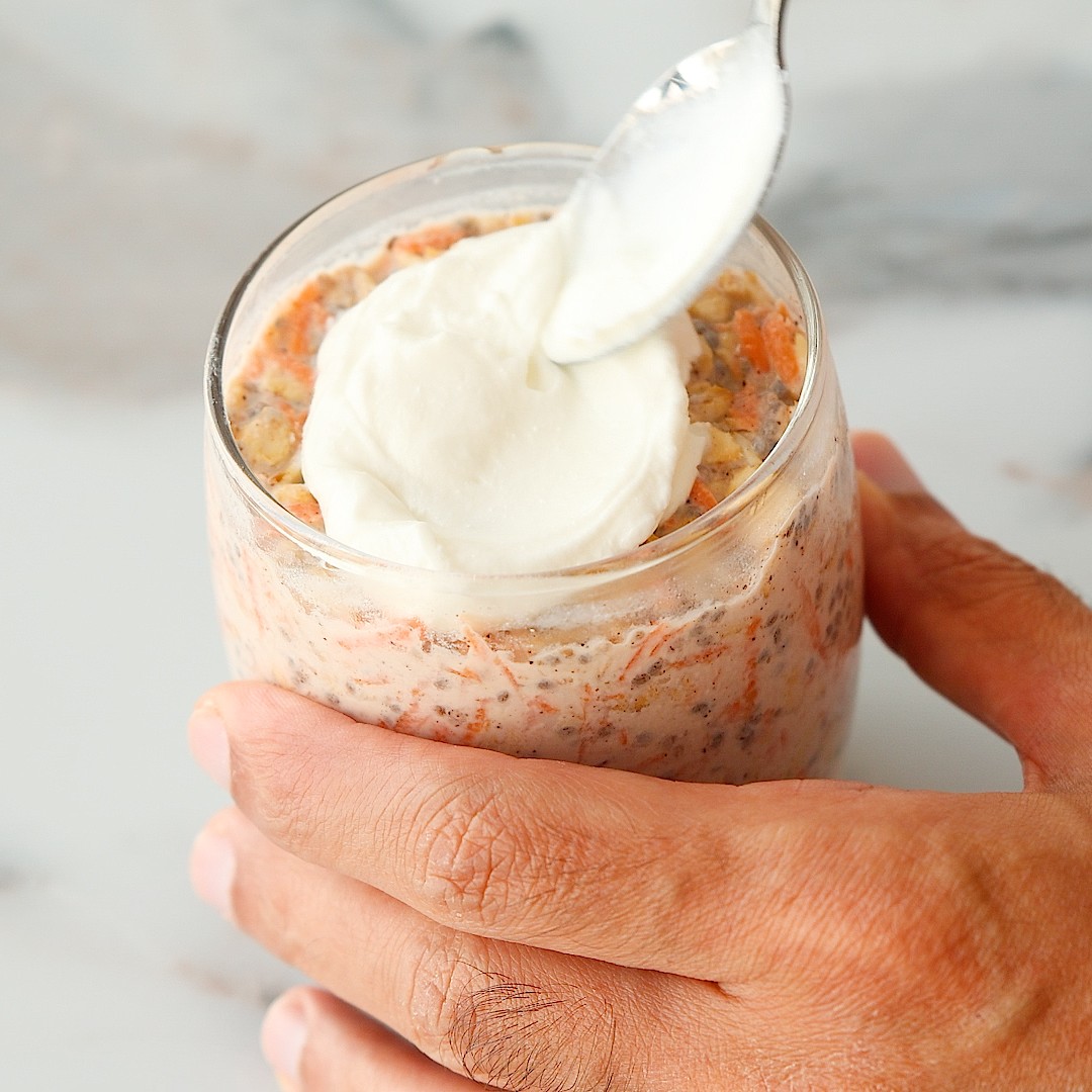 A hand holding a glass jar of carrot cake overnight oats topped with a spoonful of yogurt.