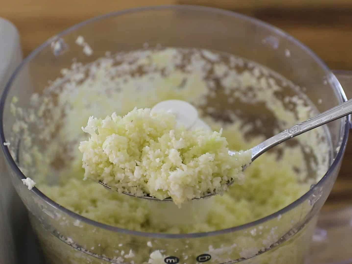 A spoon holds up freshly riced cauliflower over a food processor bowl filled with more riced cauliflower.