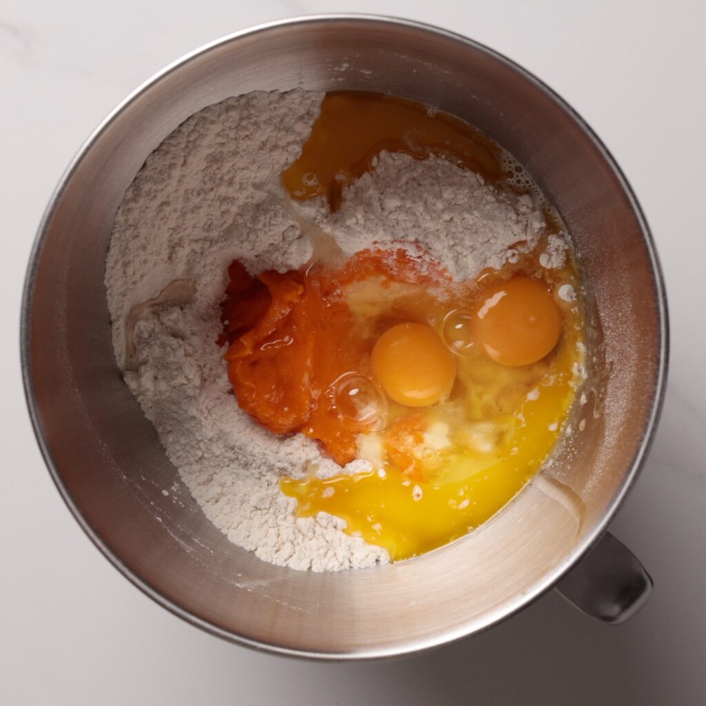 A metal mixing bowl with flour, eggs, pumpkin puree, and melted butter, ready to be mixed on a white countertop.