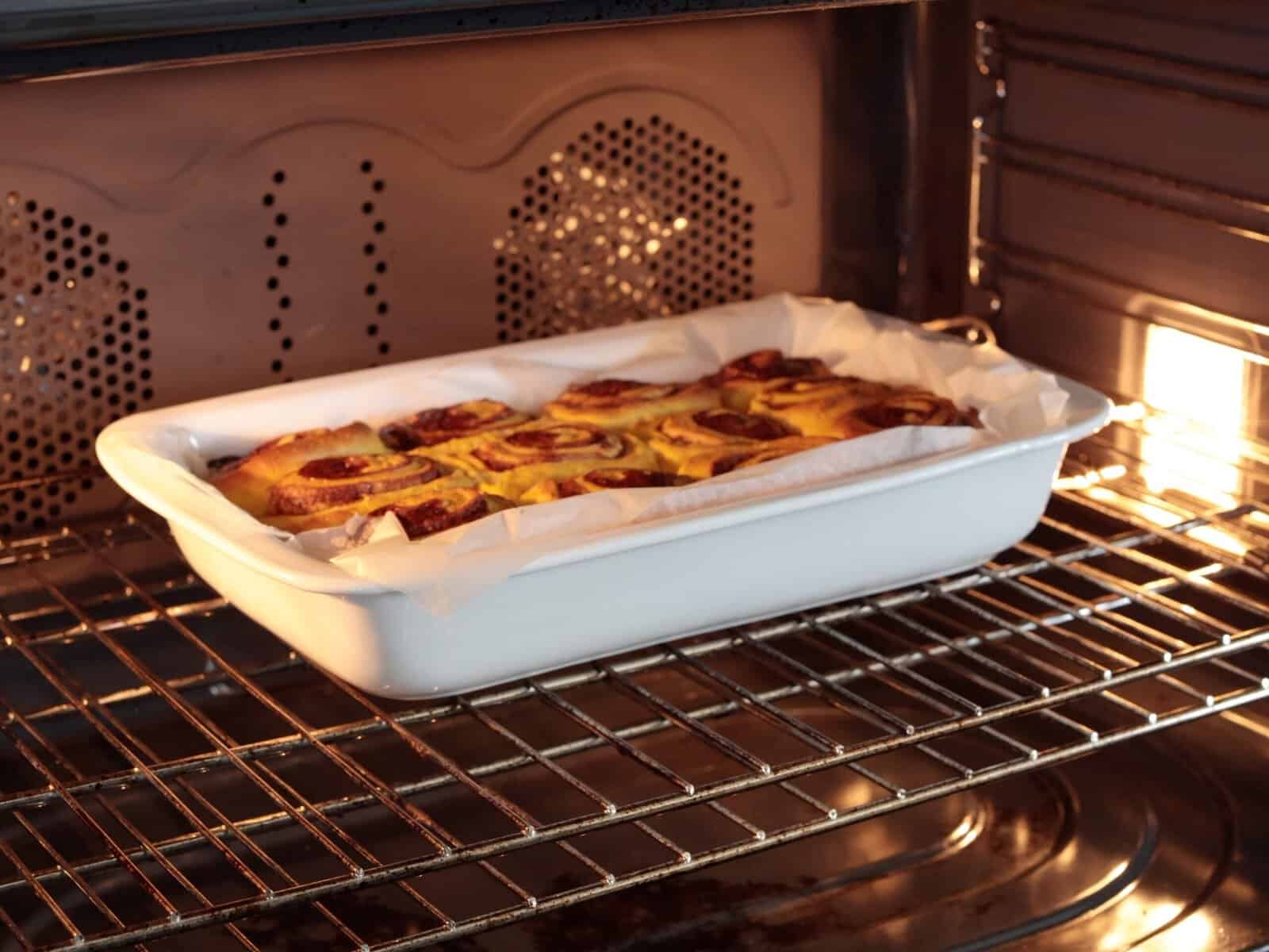 A white baking dish with cinnamon rolls, lined with parchment paper, is baking on the middle rack inside an oven, with the oven light on.