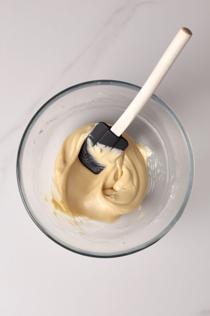 A glass bowl containing thick, creamy cream cheese frosting for pumpkin cinnamon rolls, with a black spatula, placed on a light-colored surface.