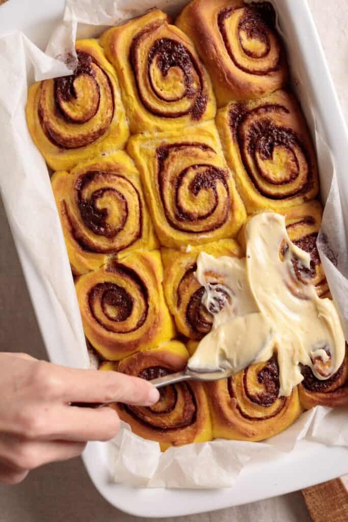 A hand spreads cream cheese frosting over freshly baked cinnamon rolls with pumpkin arranged in a white rectangular baking dish lined with parchment paper.