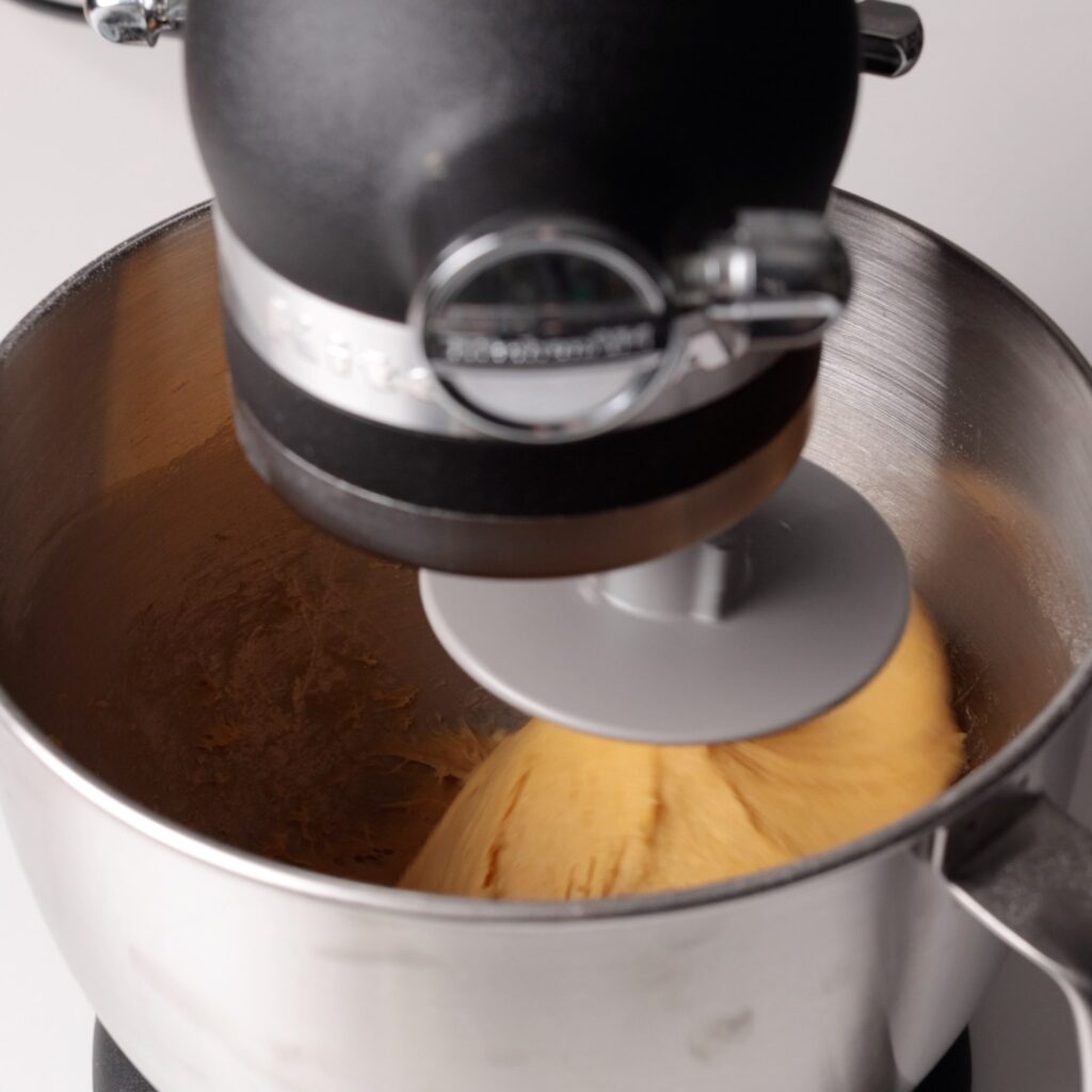 A stand mixer with a dough hook attachment is kneading dough in a stainless steel mixing bowl on a light background.