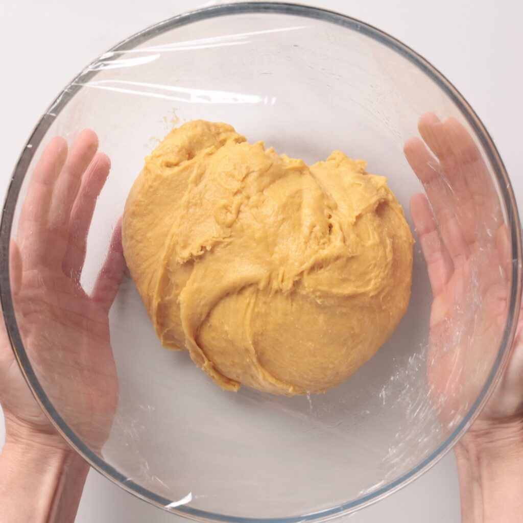 A pair of hands holds a glass bowl with a ball of yellow dough inside, covered by a layer of clear plastic wrap, against a white background.