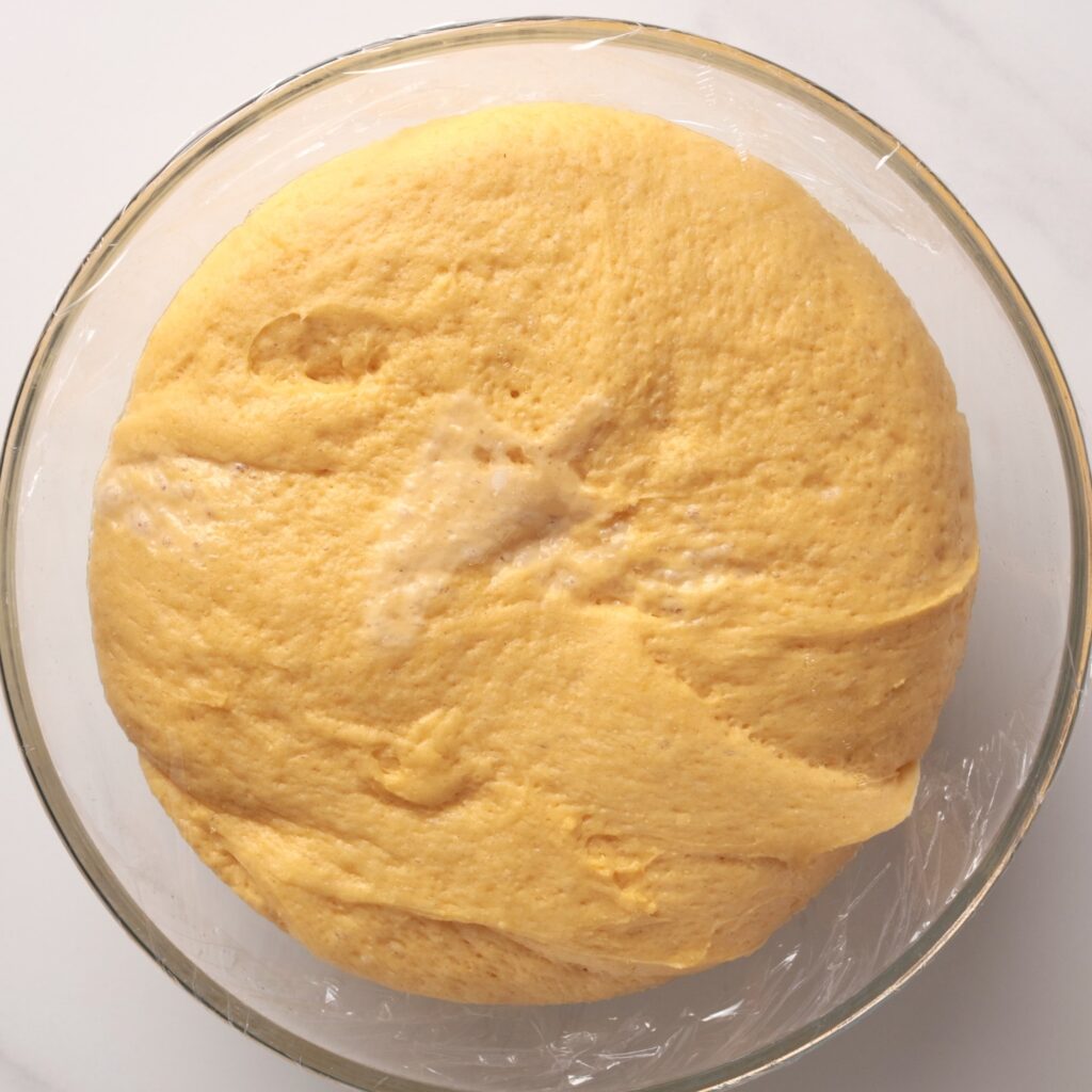A round bowl filled with risen golden dough, covered loosely with plastic wrap, sits on a white surface.