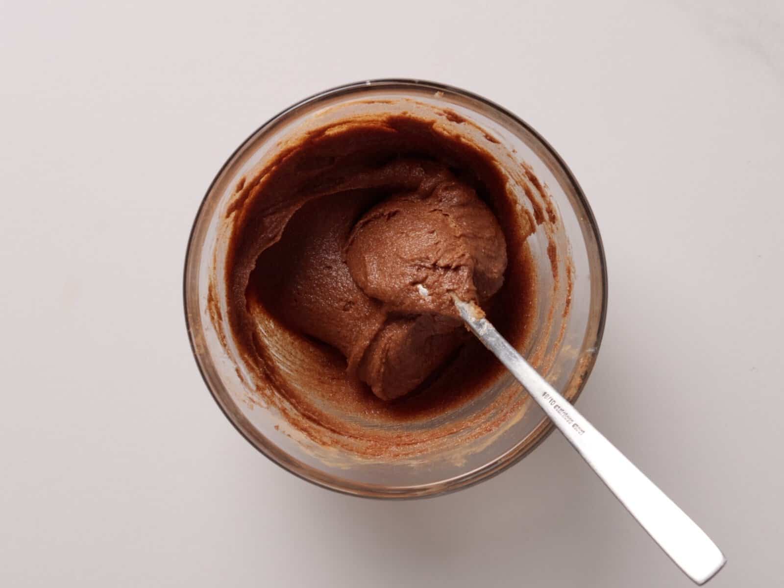 A glass bowl filled with cinnamon roll filling, with a spoon scooping some out, sits on a white surface.